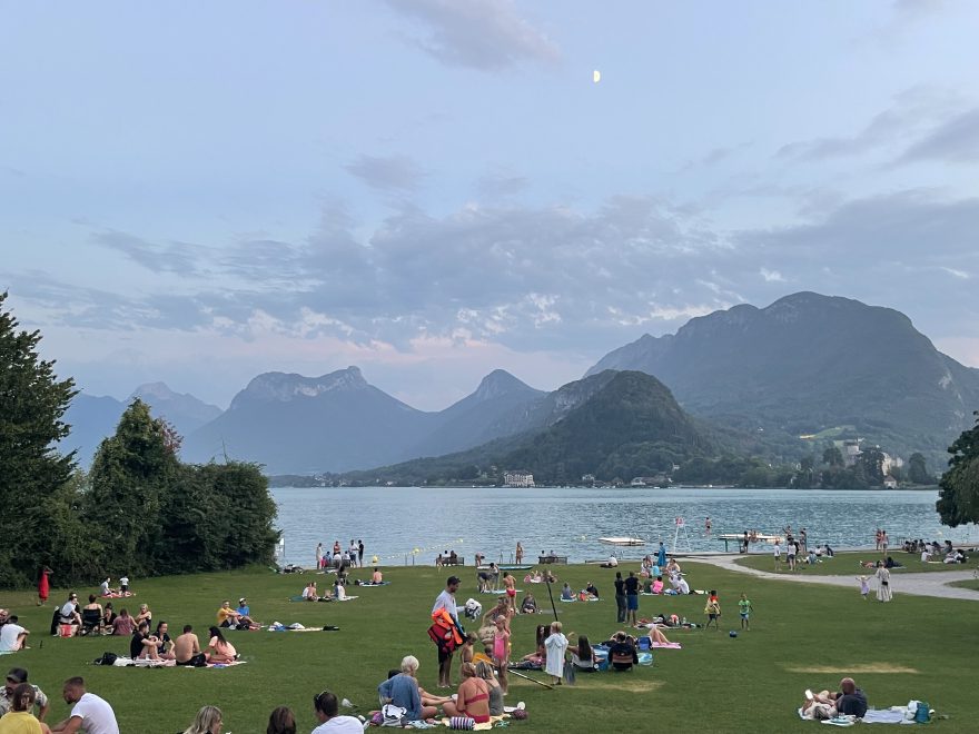 People on a lawn beside a lake with mountains in the distance